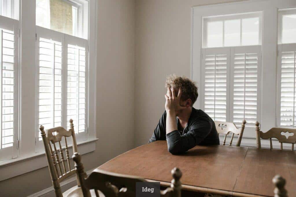 A man sits alone at a table in a bright room, displaying deep contemplation.