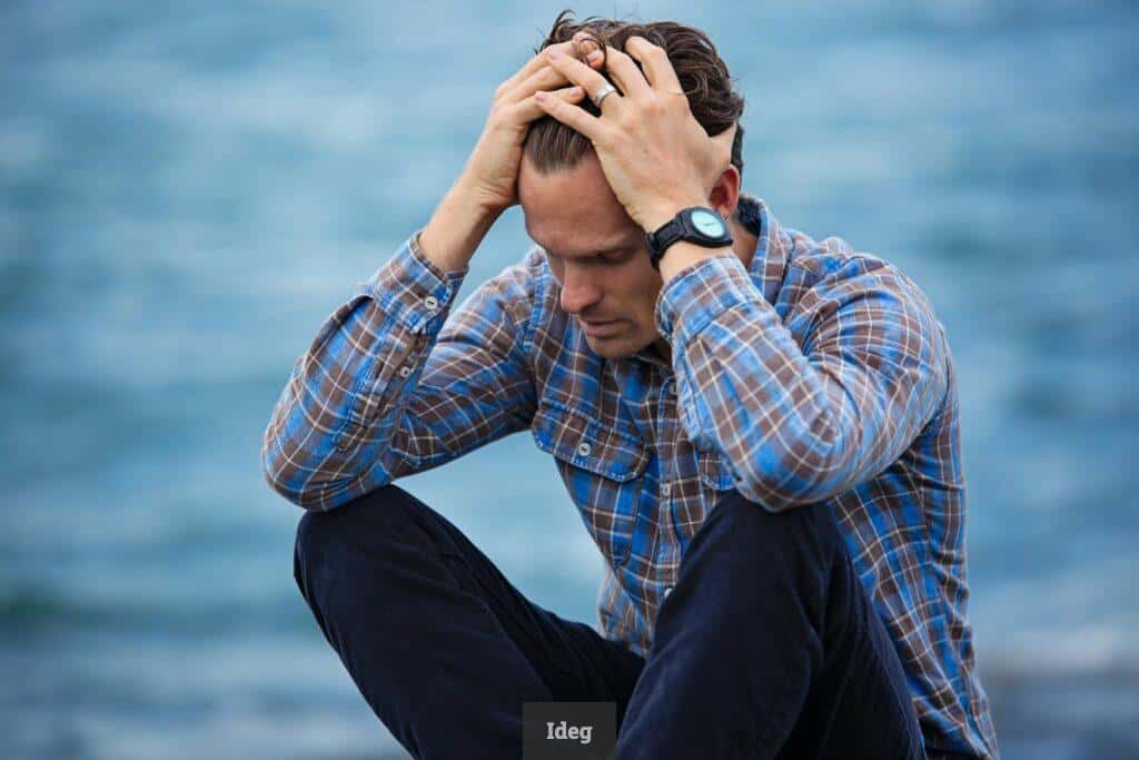 A man in a plaid shirt sits by the water looking distressed, symbolizing stress.